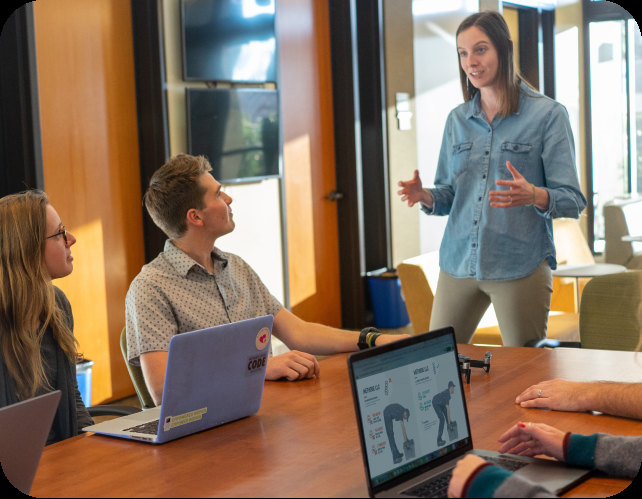 Une formatrice My Ostéo Prévention intervient dans une entreprise afin de former les collaborateurs. Quatre personnes sont assises autour d'une table avec des ordinateur et l'intervenante est debout face à eux.